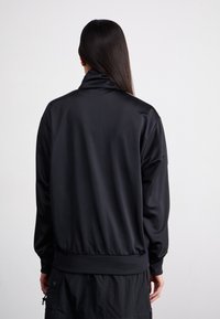 Chaqueta atlética negra con cuello alto, textura suave, puños acanalados y dobladillo ajustado. Diseñada para ofrecer comodidad y facilitar el movimiento.