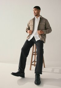 Valkoinen nappipaita päällä tanvärinen takki, mustat farkut ja mustat kengät. Malli istuu puupenkillä neutraalia taustaa vasten.