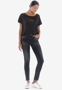 Camiseta negra de manga corta con texto en el frente, combinada con vaqueros oscuros y ajustados y zapatillas blancas. Diseño simple con un corte relajado.