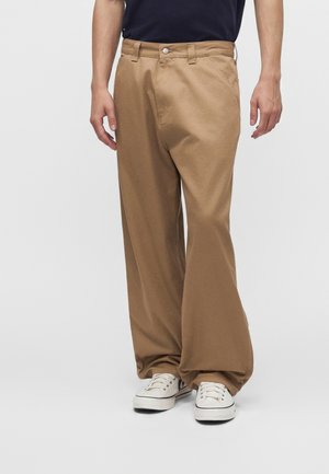 Homme portant un pantalon ample beige et des baskets blanches, debout les mains détendues le long du corps sur un fond clair uni.