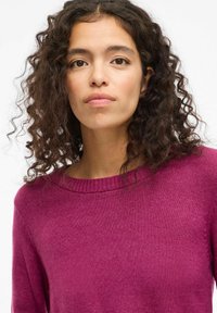 Maglione in magenta profondo a maglia, con collo rotondo e maniche lunghe. Il tessuto ha una consistenza morbida, con leggere variazioni di ombreggiatura.