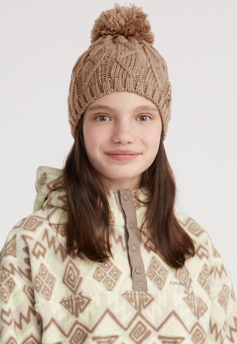Beige stickad hatt med en pom-pom på toppen, med ett strukturerat mönster. Bärs med en ljusfärgad fleecejacka prydd med geometriska mönster.