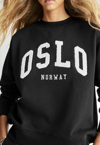 Fekete kapucnis pulóver, amelyet egy textúrázott fehér "OSLO" logó és alatta a "NORWAY" felirat díszít. Puha anyagból készült, bordázott mandzsettákkal és alsó résszel.