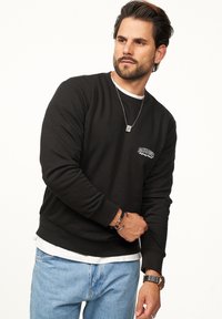 Black crewneck sweatshirt with long sleeves, textured fabric, logo on the chest, layered over a white shirt, paired with light blue jeans.