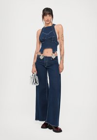 Mlada žena s tetovažama koja nosi tamnoplavu halter crop majicu i široke traperice, držeći malu srebrnu torbicu, stoji na bijeloj pozadini.