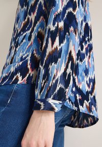 Femme portant un chemisier à manches longues avec un imprimé abstrait bleu, blanc, marron et rose, associé à un jean en denim bleu, main détendue le long du corps.