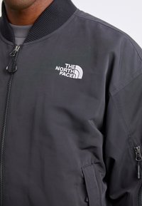Veste bomber noire avec fermeture éclair sur le devant, col côtelé et logo blanc brodé sur la poitrine gauche. Elle possède des poches latérales et une texture lisse.