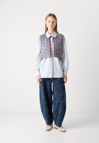 Gilet ricamato blu su una camicia colletto azzurro chiaro, abbinato a pantaloni larghi blu scuro a fantasia e ballerine beige. Design semplice e stratificato.