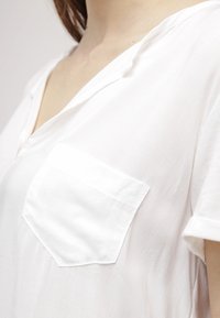 Bílá halenka s krátkým rukávem z lehké látky, s výstřihem do V a levým náprsním kapsou s jednobarevnou texturou.