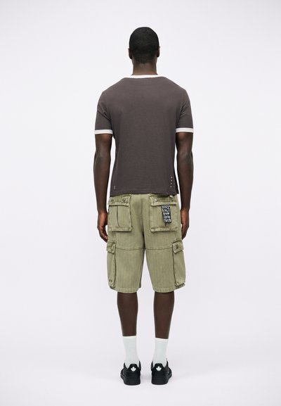 Άνδρας που κοιτάζει προς τα πίσω, φορώντας σκούρο καφέ κοντομάνικο πουκάμισο, ελιά φορτισμένα σορτς με επιθεματάκι, λευκές κάλτσες και μαύρα παπούτσια με λευκό λογότυπο φύλλου.