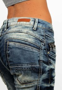 Jeans in denim blu sbiadito con cuciture a contrasto, dotati di numerose tasche e un'etichetta in pelle marrone sulla vita.