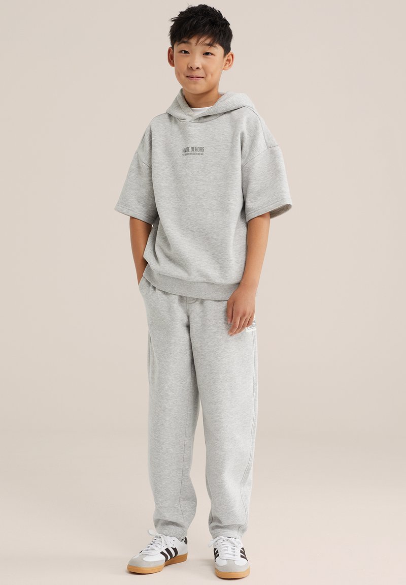 Garçon portant un sweat à capuche gris clair à manches courtes et un pantalon de jogging assorti, debout les mains dans les poches sur un fond beige.