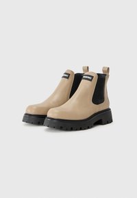 Botas de tobillo de cuero beige con paneles laterales elásticos negros, punta redonda y suela de goma negra gruesa con un patrón de tracción.