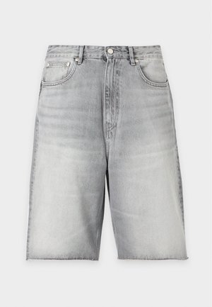 Hombre de pie con una mano en el bolsillo, vistiendo una camiseta sin mangas gris, pantalones cortos vaqueros gris claro con flecos a la altura de la rodilla y sandalias negras.