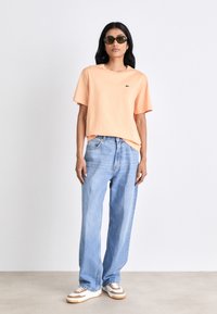 Helles oranges Baumwoll-T-Shirt mit kurzen Ärmeln und kleinem Logo, kombiniert mit locker sitzenden blauen Jeans und weißen Sneakers mit braunen Akzenten.