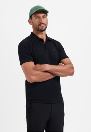 Zwarte poloshirt met een slim fit en korte mouwen, voorzien van een klein logo op de borst. Dragend met zwarte broek en een groene pet.