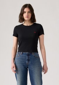 T-shirt noir court avec des manches courtes, texture côtelée, arborant un petit logo rouge "Levi's" sur la poitrine gauche. Porté avec un jean bleu taille haute.
