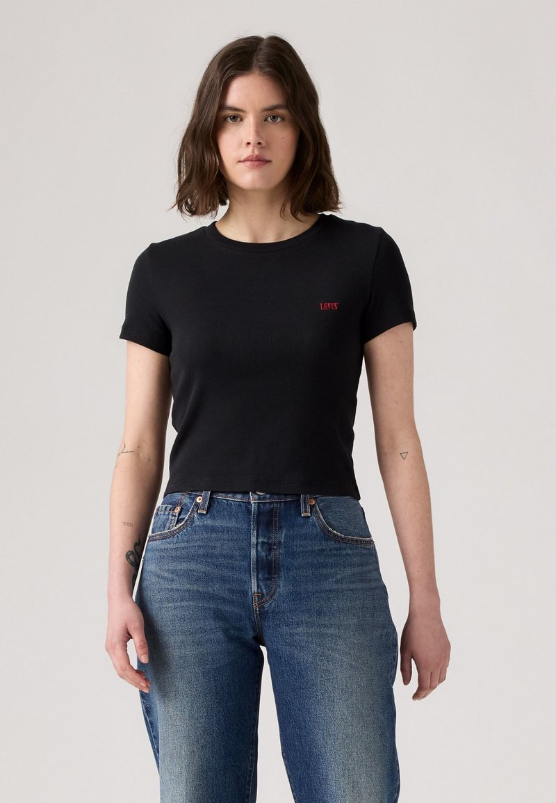 T-shirt noir court avec des manches courtes, texture côtelée, arborant un petit logo rouge "Levi's" sur la poitrine gauche. Porté avec un jean bleu taille haute.