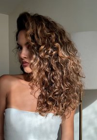 Capelli ricci, alla lunghezza delle spalle, di tonalità marrone chiaro con riflessi. Portati sciolti, con una texture definita. Modella con top bianco a coste su sfondo neutro.