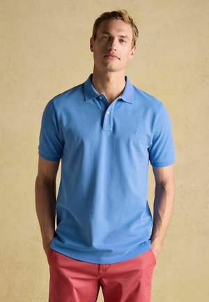 Junger Mann steht mit den Händen in den Taschen, trägt ein hellblaues Poloshirt und rosafarbene Hosen vor einem beigen Hintergrund.