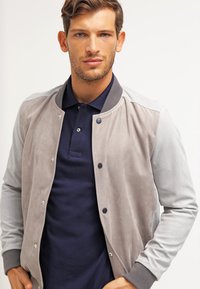 Siva nubuk bomber jakna z gladkimi rokavi, rebrastimi manžetami in gumbi spredaj, v kombinaciji z mornarsko modro polo majico spodaj.