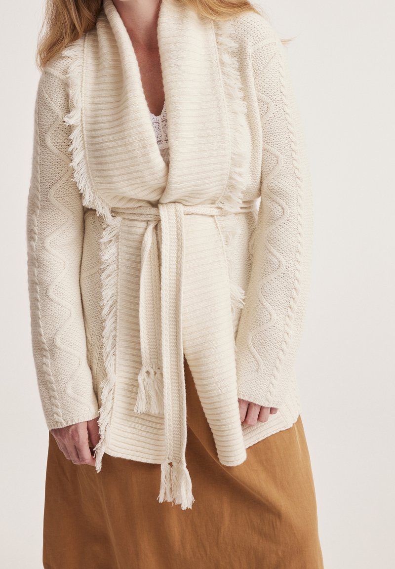 Femme portant un cardigan tricoté crème avec un col châle à franges et une ceinture, associé à une jupe marron clair.