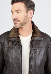 Gehaarde man met een donkere leren jas met een bontkraag over een witte wafelgebreide trui, kijkend naar links.