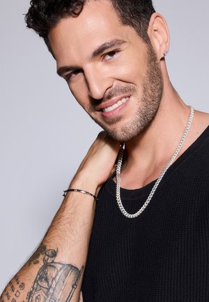 Srebrny łańcuszek z widocznymi ogniwami, noszony na czarnej dzianinowej koszulce. Na nadgarstku znajduje się prosta bransoleta, a na przedramieniu tatuaż.