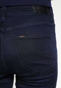Mörkblå denimjeans med en slät yta, med ett midjeband i svart läder och en liten guld "Lee"-tagg ovanför bakfickan.