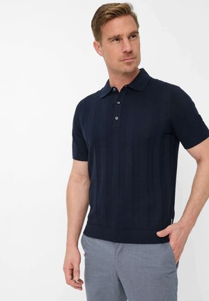 Man die een marineblauw, gebreid poloshirt met korte mouwen en lichtgrijze broek draagt, staat met één hand in zijn zak en kijkt opzij.