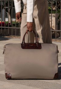 Sac de voyage beige avec des accents en cuir, doté de poignées structurées et d'une surface lisse et élégante. Fermeture éclair en haut.