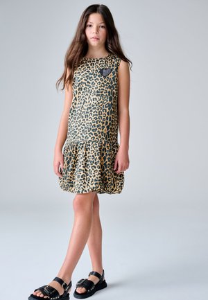 Ragazza giovane in piedi, indossa un vestito smanicato con stampa leopardata e una toppa a forma di cuore, e sandali neri con fibbie, su uno sfondo chiaro e uniforme.