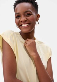 Lichtgele top met korte mouwen, van een zachte stof en subtiele naden. Het model glimlacht en draagt zilveren creolen.