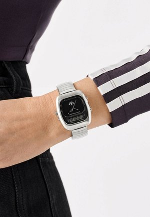 Montre numérique carrée en argent avec un cadran noir, présentant un design minimaliste, bracelet en métal avec des accents texturés, portée au poignet.