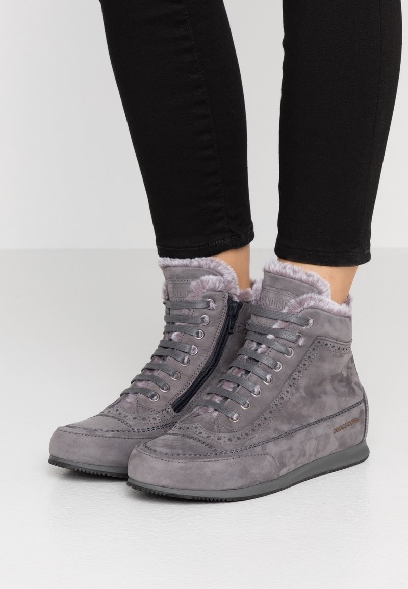 Personne portant des baskets montantes en daim gris avec doublure en fourrure, lacets et fermetures éclair latérales, associées à un pantalon noir ajusté.