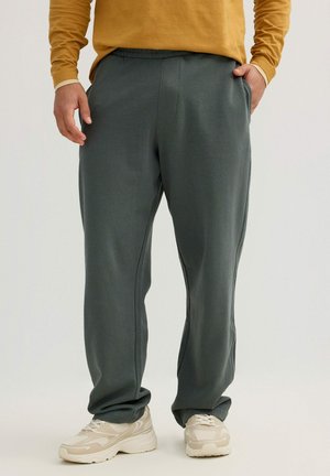 Hombre con pantalones de chándal gris oscuro, zapatillas beige y blancas, y una camiseta de manga larga mostaza con una mano en el bolsillo.