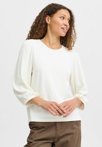 Weißer strukturierter Pullover mit Rundhalsausschnitt und Puffärmeln. Der Stoff zeigt ein dezentes Rippmuster und wird mit braunen Hosen kombiniert.