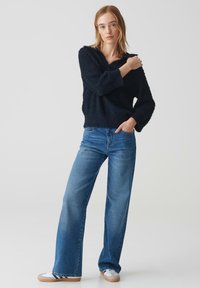 Pull en laine bleu marine avec un col en V et des manches larges, associé à un jean flare en denim bleu taille haute et des baskets blanches avec des semelles beiges.