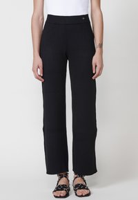 Pantalons en tricot côtelé noirs avec une taille haute, présentant un ajustement serré en haut et un ourlet évasé. Associés à des sandales noires à lanières.