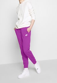 Violet sweatpants dengan cuff elastis dan saku samping, dipadukan dengan hoodie beige dan sepatu sneakers putih. Dilengkapi dengan logo Nike di celana.