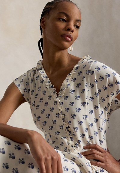 Nainen, jolla on letitetyt hiukset ja valkoinen mekko, jossa on pieni sininen kukkakuvio ja röyhelöinen kaulus, poseeraa käsi lantiolla yksiväristä taustaa vasten.