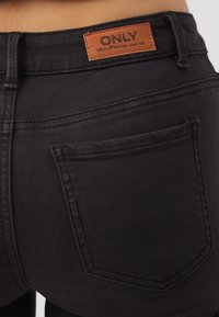 Svarta denimjeans med standardpassform, med ett brunt lädermärke och traditionella bakfickor. Slät textur, synlig sömnad.