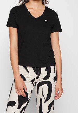 T-shirt noir en V fabriqué en coton doux, avec des manches courtes et un petit logo accentué, associé à un pantalon crème présentant un motif noir audacieux.