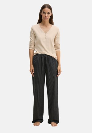 Vrouw staat blootsvoets en draagt een beige henley-top met lange mouwen en een losse zwarte broek met witte verticale krijtstrepen.