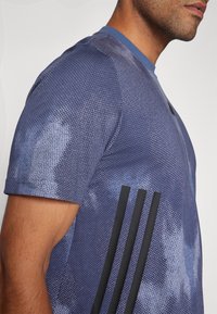 Man som bär en blåmönstrad kortärmad sportskjorta med tre svarta vertikala ränder på sidan och en rund halsringning.