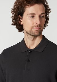 Černé polo tričko se strukturovanou tkaninou, zapínáním na dva knoflíky a krátkým límečkem. Detailní pohled zvýrazňující strukturu tkaniny.