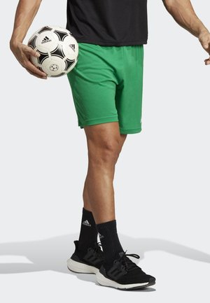 Groene sportshorts van lichtgewicht materiaal, gecombineerd met zwarte sneakers en sokken met het Adidas-logo, terwijl een voetbal wordt vastgehouden.