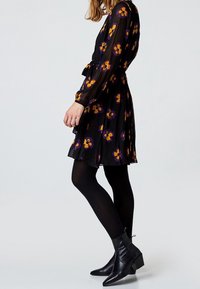Robe noire à manches longues avec un motif floral en violet et orange. La jupe est plissée, associée à des collants opaques noirs et des bottes de cheville.