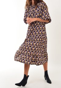 Vestido midi de estampado púrpura y amarillo con un dobladillo en capas, mangas abullonadas y escote fruncido. Lleva botas negras hasta el tobillo.