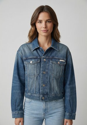 Kvinde med skulderlangt brunt hår iført en blå denimjakke over lyseblå jeans, vendt mod kameraet foran en ensfarvet grå baggrund.